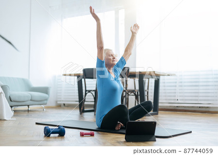 elderly woman sitting in a yoga pose in front of an open laptop. elderly woman sitting in a yoga pose in front of an open laptop. 87744059