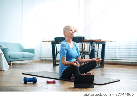 elderly woman sitting in a yoga pose in front of an open laptop. elderly woman sitting in a yoga pose in front of an open laptop. 87744060