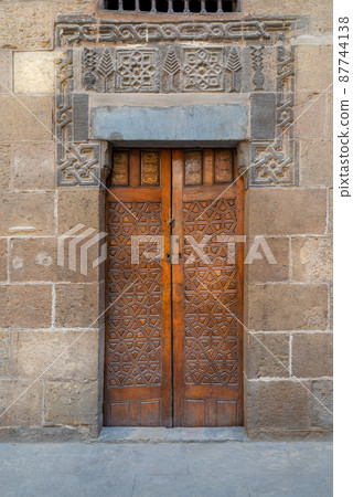 Wooden ornate door with geometrical engraved patterns on external old decorated bricks stone wall Wooden ornate door with geometrical engraved patterns on external old decorated bricks stone wall 87744138