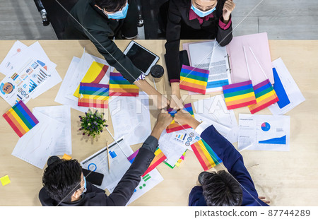 top view Successful workers Group of asian business partners wearing protective mask casual with diverse genders LGBT putting their hands together with Paper LGBT Flag at it at meeting in room office 87744289