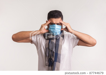 Unhealthy man in surgical mask and scarf suffering headache, rubbing painful temples, having symptoms of flu, contagious coronavirus infection 2019-nCoV. studio shot isolated on white background 87744679