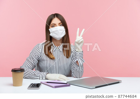 Optimistic woman employee sitting safe healthy with protective mask and gloves during quarantine, showing victory, peace gesture. Working at home office in self-isolation, coronavirus outbreak. indoor Optimistic woman employee sitting safe healthy with protective mask and gloves during quarantine, showing victory, peace gesture. Working at home office in self-isolation, coronavirus outbreak. indoor 87744684