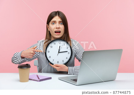 Nervous anxious woman office worker pointing finger at big wall clock in her hands sitting at workplace, working late hours, overtime job. Indoor studio shot isolated on pink background 87744875