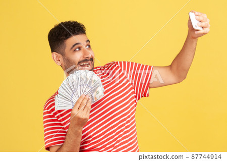 Happy positive man with beard in striped t-shirt holding and showing on smartphone camera fan of hundred dollars bills, blogger showing his income. Indoor studio shot isolated on yellow background Happy positive man with beard in striped t-shirt holding and showing on smartphone camera fan of hundred dollars bills, blogger showing his income. Indoor studio shot isolated on yellow background 87744914