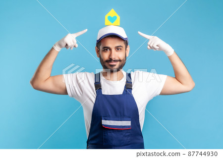 Portrait of positive handyman, mover guy in workwear and safety gloves pointing paper house on head. Home repair advertisement, maintenance or moving services, help in relocation. studio shot isolated Portrait of positive handyman, mover guy in workwear and safety gloves pointing paper house on head. Home repair advertisement, maintenance or moving services, help in relocation. studio shot isolated 87744930