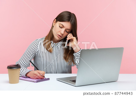 Diligent secretary in striped shirt receiving calls on phone and making notes, writing down information, sitting at workplace with laptop. Indoor studio shot isolated on pink background 87744943