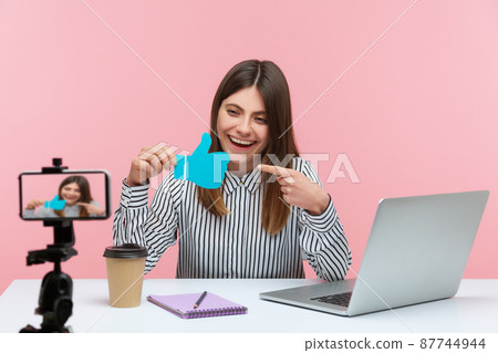 Smiling satisfied woman blogger pointing finger at blue thumbs up template asking to like her posts, streaming on smartphone camera. Indoor studio shot isolated on pink background Smiling satisfied woman blogger pointing finger at blue thumbs up template asking to like her posts, streaming on smartphone camera. Indoor studio shot isolated on pink background 87744944