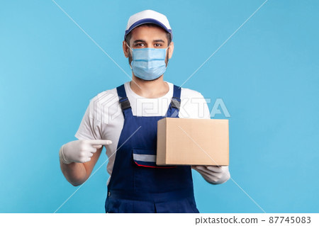 Cargo transportation, delivery service. Friendly worker in uniform and mask pointing at cardboard box and smiling to camera. Courier carrying parcel. Online express shipping, relocation service. 87745083