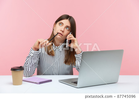 Dreamy thoughtful woman office worker sitting at workplace with laptop, contemplating and making notes at notepad talking phone with client. Indoor studio shot isolated on pink background 87745096