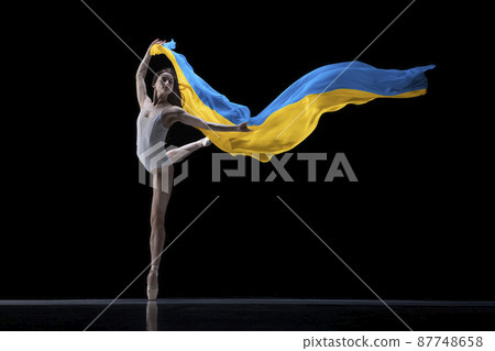 Young graceful classic ballerina dancing with cloth painted in blue and yellow colors of Ukraine flag on dark studio background. Art, peace, freedom and rights 87748658