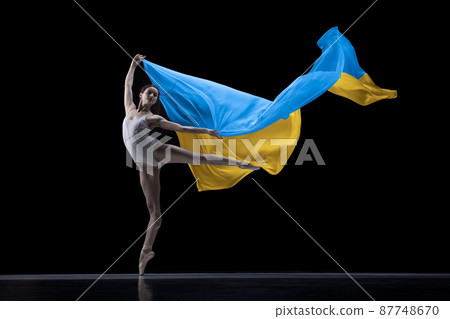 Young graceful classic ballerina dancing with cloth painted in blue and yellow colors of Ukraine flag on dark studio background. Art, peace, freedom and rights 87748670
