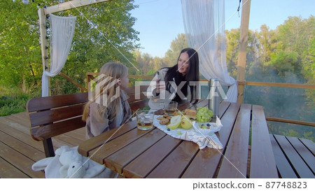 Mom and daughter are sitting at a wooden table in nature. A mother takes a picture of her daughter on her phone. Family on a picnic. 87748823