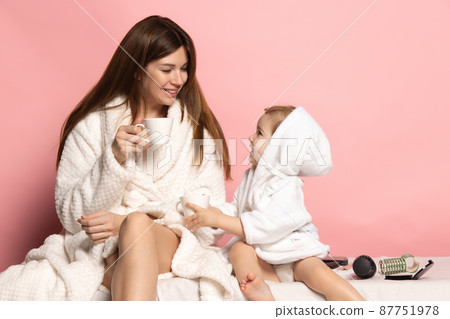 Young beautiful happy woman and little kid, mother and daughter wearing bathrobe drinking coffee isolated on pink background. Mother's Day, family, childhood 87751978