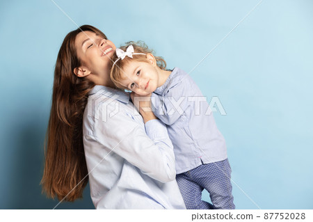 Studio shot of beautiful happy woman and little kid, mother and daughter isolated on blue background. Mother's Day celebration. Concept of family, childhood Studio shot of beautiful happy woman and little kid, mother and daughter isolated on blue background. Mother's Day celebration. Concept of family, childhood 87752028