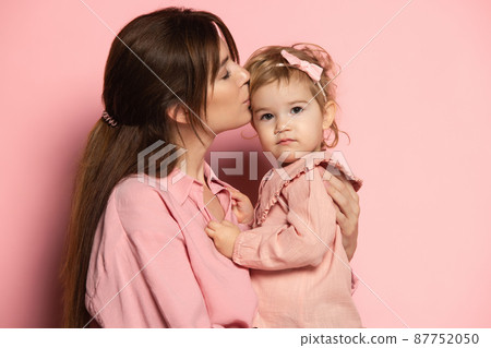 Kiss. Portrait of young woman and little girl, mother and daughter isolated on pink studio background. Mother's Day celebration. Concept of family, childhood, motherhood Kiss. Portrait of young woman and little girl, mother and daughter isolated on pink studio background. Mother's Day celebration. Concept of family, childhood, motherhood 87752050