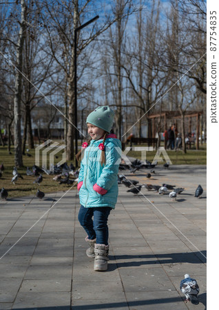 Preschool-aged girl in a blue jacket twirls around herself in front of a flock of city doves. Portrait of a five-year-old child with long pigtails holding her hands in her pockets. Sunny spring day. Preschool-aged girl in a blue jacket twirls around herself in front of a flock of city doves. Portrait of a five-year-old child with long pigtails holding her hands in her pockets. Sunny spring day. 87754835