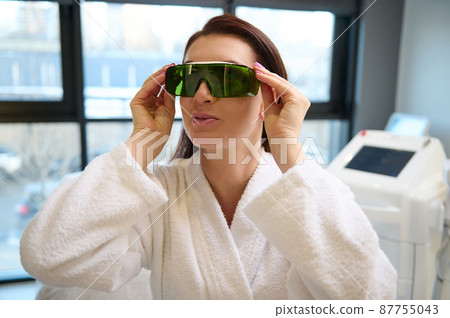 Confident Caucasian middle aged woman putting on protective UV eyeglasses, ready for beauty treatment with laser apparatus in a wellness spa centre with contemporary medical equipments Confident Caucasian middle aged woman putting on protective UV eyeglasses, ready for beauty treatment with laser apparatus in a wellness spa centre with contemporary medical equipments 87755043