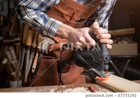 Carpenter is sawing a wood plank with electric jig saw machine in carpentry workshop. Workwood DIY 87755292