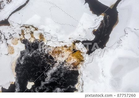 Top view of the river waterfall. Winter aerial view of a freezing river. Animal tracks on the ice covered with snow. Top view of the river waterfall. Winter aerial view of a freezing river. Animal tracks on the ice covered with snow. 87757260