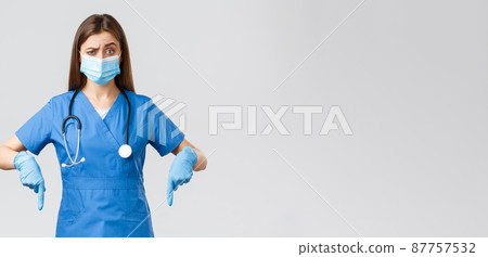 Covid-19, preventing virus, health, healthcare workers and quarantine concept. Skeptical and unsure female nurse or doctor in blue scrubs, personal protective equipment, pointing fingers down unsure 87757532