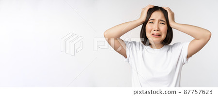 Desperate young korean woman holding hands on head, panicking, crying and standing distressed against white background 87757623