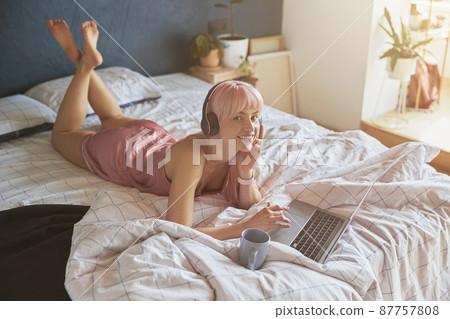 Pink haired woman in silk pajamas rests using laptop and headphones on large bed Pink haired woman in silk pajamas rests using laptop and headphones on large bed 87757808