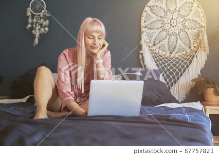 Lady with long dyed pink hair surfs internet with laptop resting on bed Lady with long dyed pink hair surfs internet with laptop resting on bed 87757821
