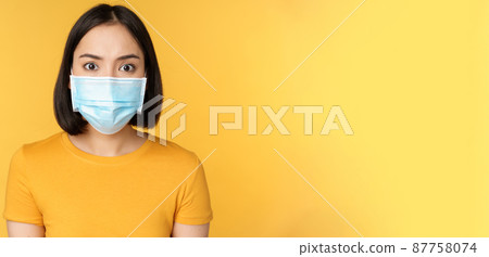 Portrait of shocked asian woman looking concerned and startled at camera, wearing covid-19 medical face mask, standing against yellow background Portrait of shocked asian woman looking concerned and startled at camera, wearing covid-19 medical face mask, standing against yellow background 87758074