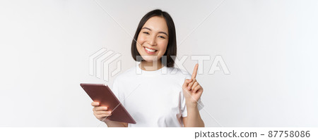 Enthusiastic asian woman with tablet, raising finger and looking amazed, pointing up, standing over white background 87758086