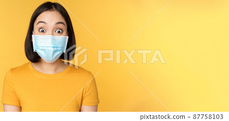 Covid-19 and medical concept. Close up portrait of asian woman in face mask, looking surprised and amazed at news, standing over yellow background Covid-19 and medical concept. Close up portrait of asian woman in face mask, looking surprised and amazed at news, standing over yellow background 87758103