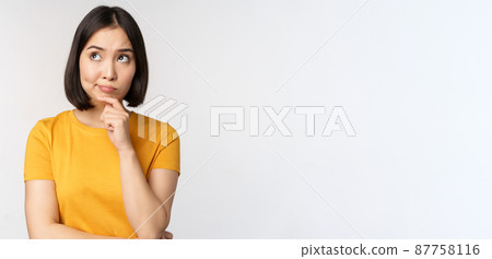Image of thinking asian woman, looking aside and pondering, making decision, standing in yellow tshirt over white background 87758116
