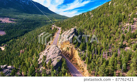 Road cutting through large mountain rock in valley of pine trees Road cutting through large mountain rock in valley of pine trees 87762421