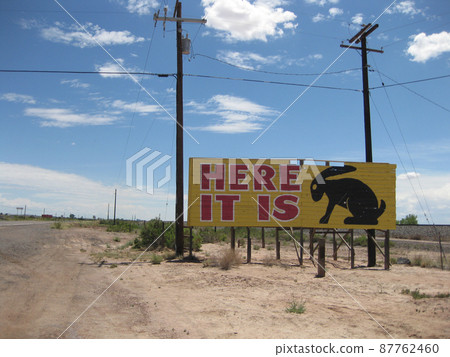Along a desert road, a yellow billboard sign reads Here It Is with a rabbit's silhouette Along a desert road, a yellow billboard sign reads Here It Is with a rabbit's silhouette 87762460