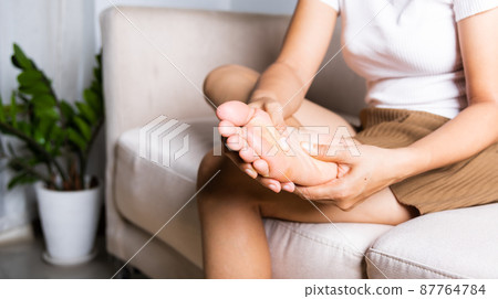 Asian woman sitting on sofa feeling pain in her foot at home 87764784