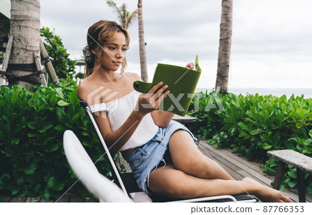 Young woman reading ebook in tropical yard 87766353
