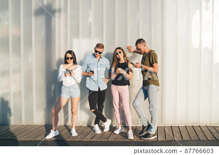 Group of confident friends standing near wall and browsing smartphones Group of confident friends standing near wall and browsing smartphones 87766603