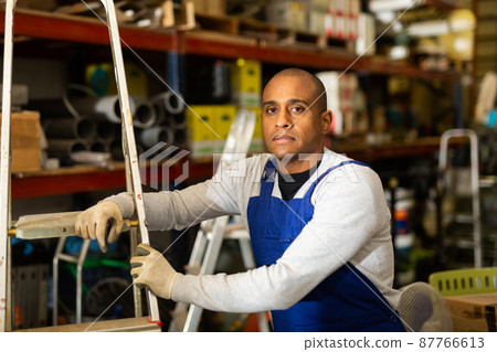 Positive warehouse worker stands next to stepladder and tool shelves in hardware store Positive warehouse worker stands next to stepladder and tool shelves in hardware store 87766613