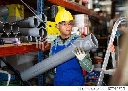 Plumber in protective helmet and overalls selects plastic pipes in hardware store 87766735