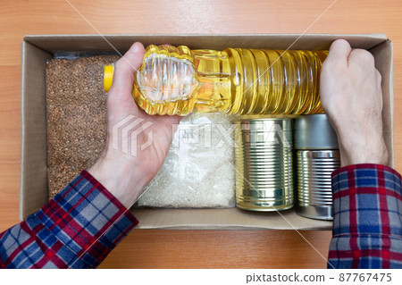 Man in a shirt stacks food in case of emergency, natural disasters, self-isolation, pandemics, covid 19. Food with a long shelf life. Canned food, oil, rice, buckwheat. Help for the starving 87767475