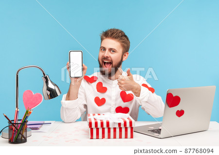 Smiling satisfied man with beard in white shirt covered with sticky hearts showing thumbs up holding smartphone with empty display. Indoor studio shot isolated on blue background 87768094