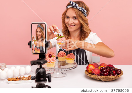 Food vlogger holding small cake decorating with cream and cherry, looking at camera on smart phone on tripod and smiling, recording video for vlog. Indoor studio shot isolated on pink background. 87768096
