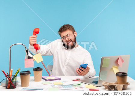 Busy overworked man sitting at workplace all covered with sticky notes and holding three phones in hands, answering calls, service center. Indoor studio shot isolated on blue background. Busy overworked man sitting at workplace all covered with sticky notes and holding three phones in hands, answering calls, service center. Indoor studio shot isolated on blue background. 87768105
