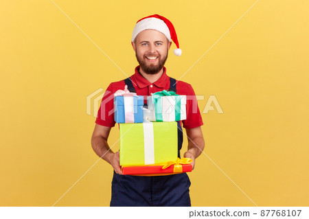 Positive smiling delivery man in blue uniform red t-shirt and santa claus new year hat on head holding many gift boxes, delivery of holiday presents. Indoor studio shot isolated on yellow background Positive smiling delivery man in blue uniform red t-shirt and santa claus new year hat on head holding many gift boxes, delivery of holiday presents. Indoor studio shot isolated on yellow background 87768107