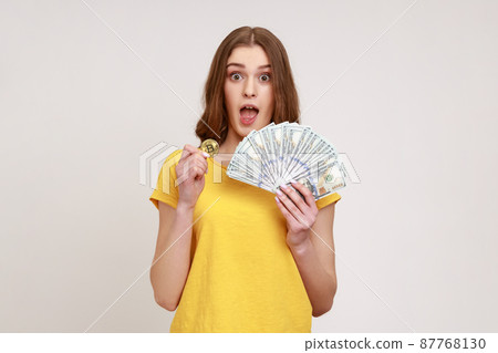 Excited beautiful teen girl in yellow T-shirt holding in hands and showing dollar bills and golden coin of crypto currency, shocked by value of bitcoin. Indoor studio shot isolated on gray background. Excited beautiful teen girl in yellow T-shirt holding in hands and showing dollar bills and golden coin of crypto currency, shocked by value of bitcoin. Indoor studio shot isolated on gray background. 87768130
