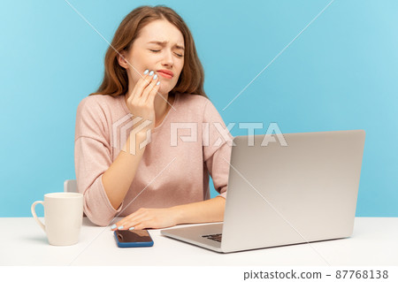 Unhealthy young woman employee sitting at workplace with laptop and holding painful cheek, suffering intense toothache, inflamed gums, dental problems. indoor studio shot isolated on blue background Unhealthy young woman employee sitting at workplace with laptop and holding painful cheek, suffering intense toothache, inflamed gums, dental problems. indoor studio shot isolated on blue background 87768138