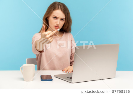 Impolite furious young woman employee sitting at workplace with laptop, showing middle finger, expressing disrespect and hate, conflict with coworkers. indoor studio shot isolated on blue background 87768156
