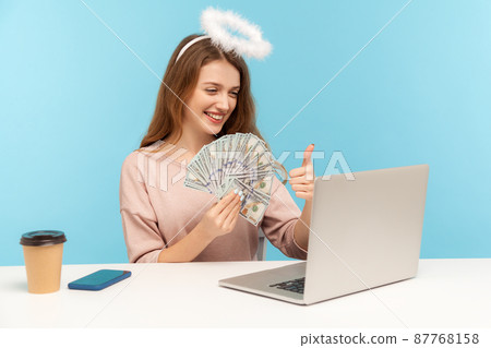 Recommending big money profit. Lovely angelic rich woman with nimbus holding dollar banknotes and showing thumbs up while talking video call on laptop. indoor studio shot isolated on blue background Recommending big money profit. Lovely angelic rich woman with nimbus holding dollar banknotes and showing thumbs up while talking video call on laptop. indoor studio shot isolated on blue background 87768158
