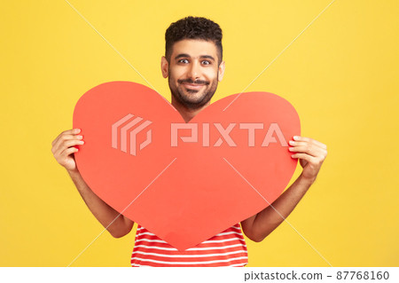 Positive smiling man in love holding in hands big red paper heart looking at camera with smile, confessing his love, romantic relationships. Indoor studio shot isolated on yellow background Positive smiling man in love holding in hands big red paper heart looking at camera with smile, confessing his love, romantic relationships. Indoor studio shot isolated on yellow background 87768160