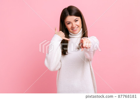 Smiling positive young adult female showing call me gesture with hand and pointing finger at camera, wearing white casual style sweater. Indoor studio shot isolated on pink background. 87768181