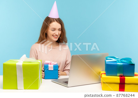 Funny cheerful woman with party cone sitting at workplace surrounded by many presents and talking on video call, accepting congratulations via online webcam communication. indoor studio shot, isolated Funny cheerful woman with party cone sitting at workplace surrounded by many presents and talking on video call, accepting congratulations via online webcam communication. indoor studio shot, isolated 87768244